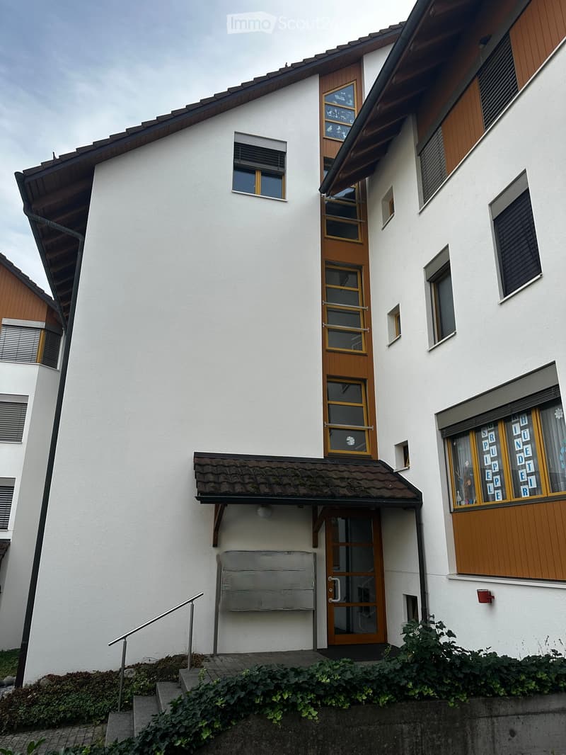 Two-story house with white exterior, wooden doors and windows, small porch with steps, and greenery at the base.