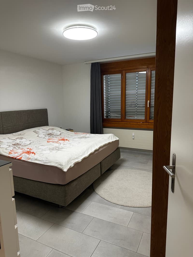 Modern bedroom with a large bed, floral duvet, nightstand, and large window with wooden frame and blinds.
