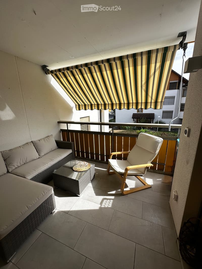 Balcony with a striped awning, a wicker sofa, a wooden chair, and a small table with a cushion.