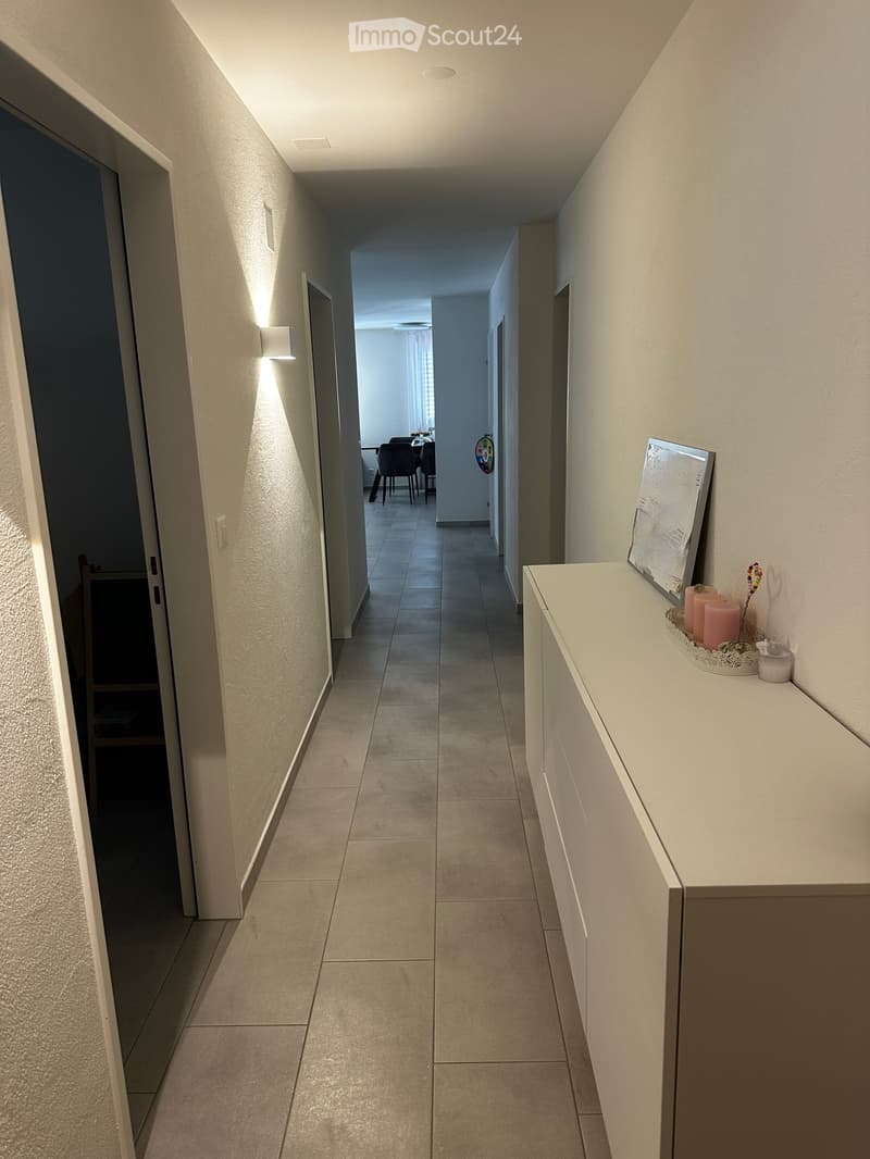 Clean hallway with tiled floor, white walls, and modern lighting fixtures. A small console table with candles and a framed picture is visible on the right side.