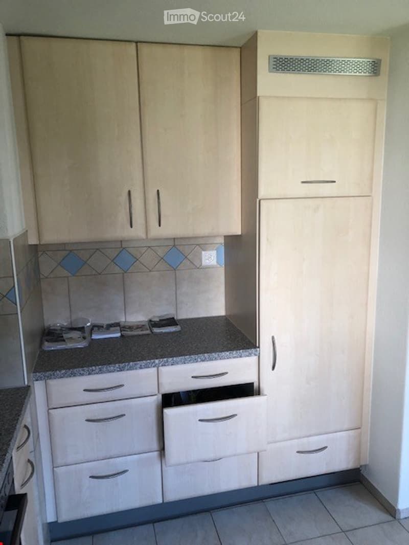 Light wood kitchen with light-colored cabinets, drawers, and a granite countertop. Blue mosaic tiles are visible on the backsplash.
