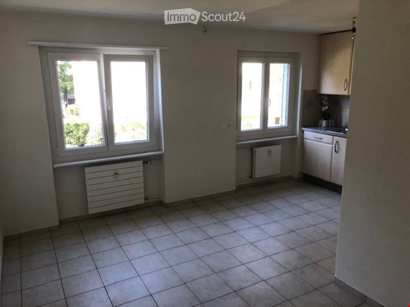 Empty room with tiled floor, two windows, radiator, and a small kitchen area with cabinets and sink.