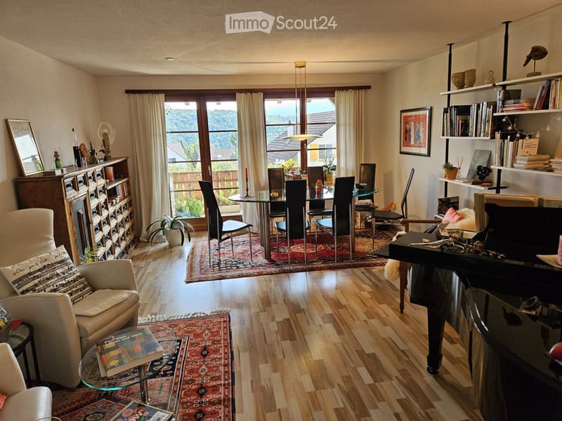 Spacious living and dining area with wooden flooring, large windows, and a view of greenery. Features a sofa, armchair, coffee table, dining table with chairs, bookshelf, piano, and decorative items.