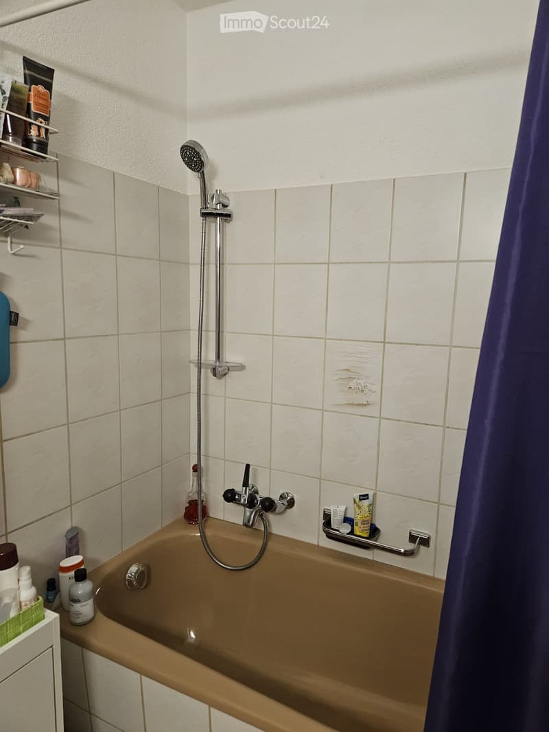 Bathtub with a handheld showerhead and faucet, surrounded by tiled walls and a shelf holding various toiletries.