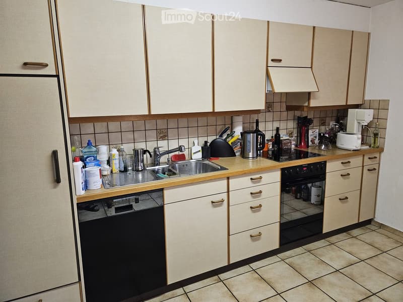 The kitchen features light-colored cabinets, a tiled backsplash, and various appliances including a sink, oven, dishwasher, coffee maker, and toaster. Cleaning supplies and bottles are placed on the counter.