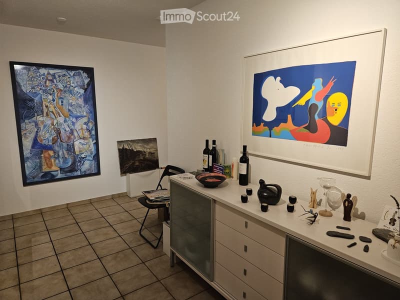 A modern gallery or display room with a white table holding various art pieces, bottles, and decorative items, framed artworks on the walls, and a chair near the table.