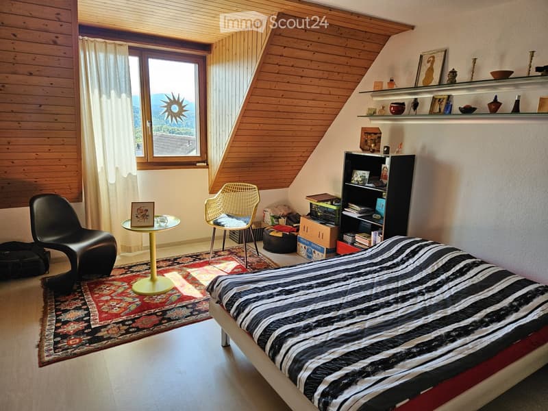 Cozy attic bedroom with wooden slanted ceilings, large window with view, bed with striped bedding, wicker chair, small round table, shelves with decorative items, and patterned rug.