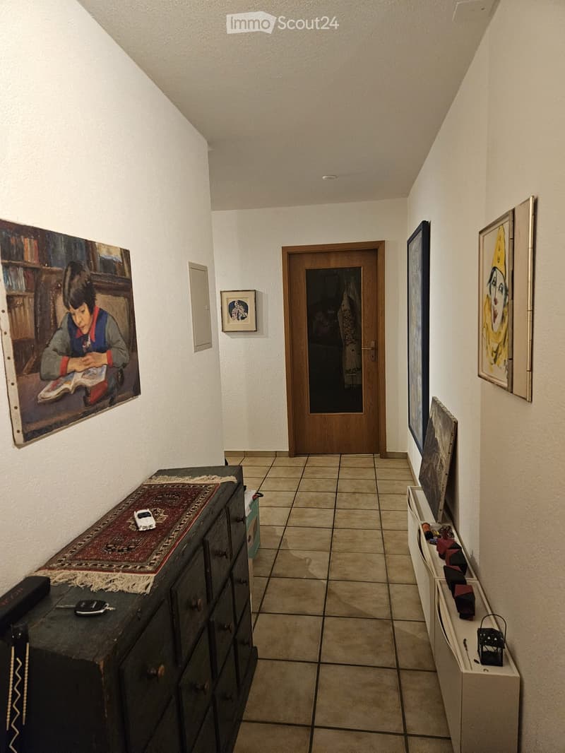 A narrow hallway with tiled flooring, white walls, and several framed artworks displayed on the walls. A wooden door is visible at the end of the hallway, and a small table with a rug and some items is placed on the left side.