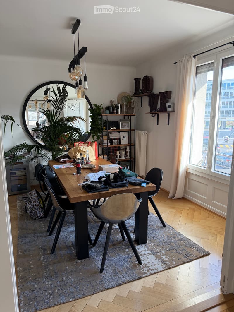 A modern, well-lit room with wooden flooring, a large round mirror, a wooden dining table with chairs, a shelf with books and decorative items, and a large window with curtains.