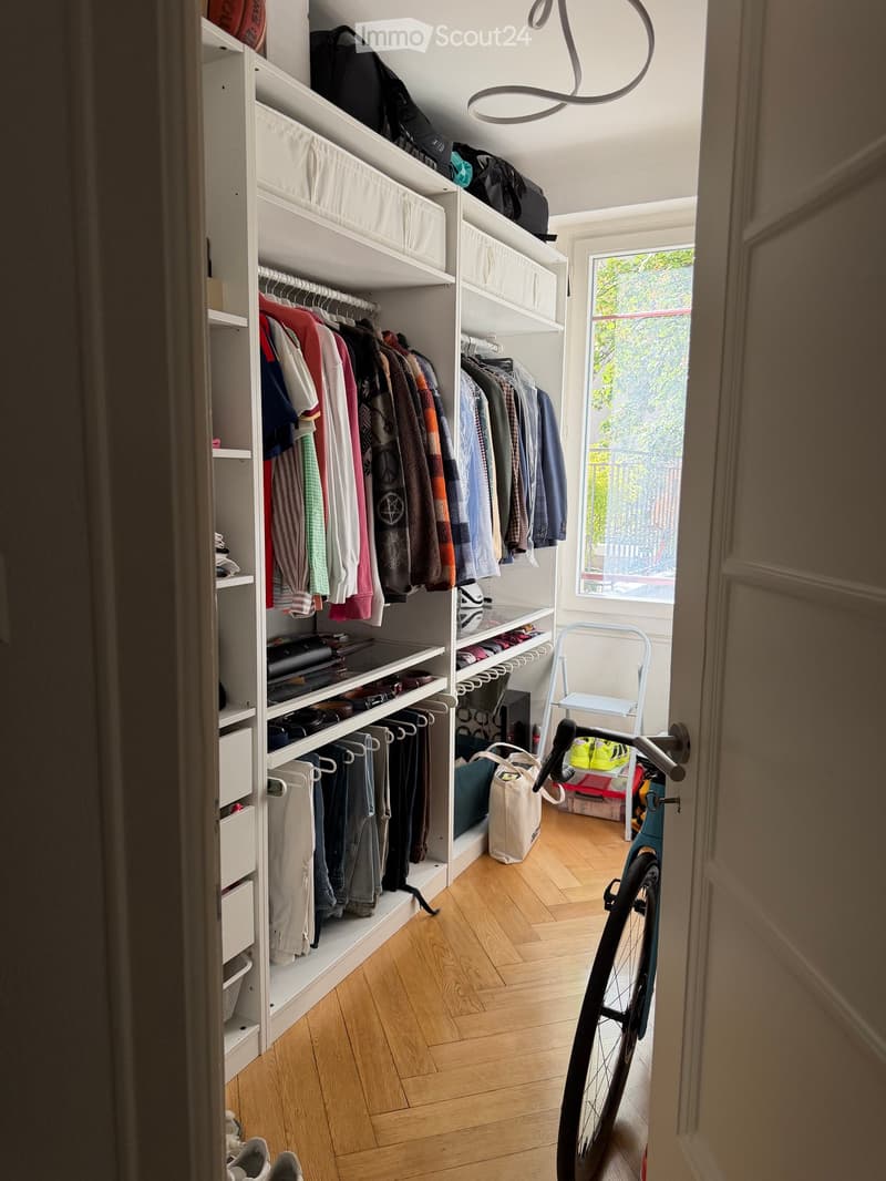A well-organized walk-in closet with white built-in wardrobes, wooden flooring, and a bicycle leaning against the wall. The closet contains neatly arranged clothing, shoes, and accessories.