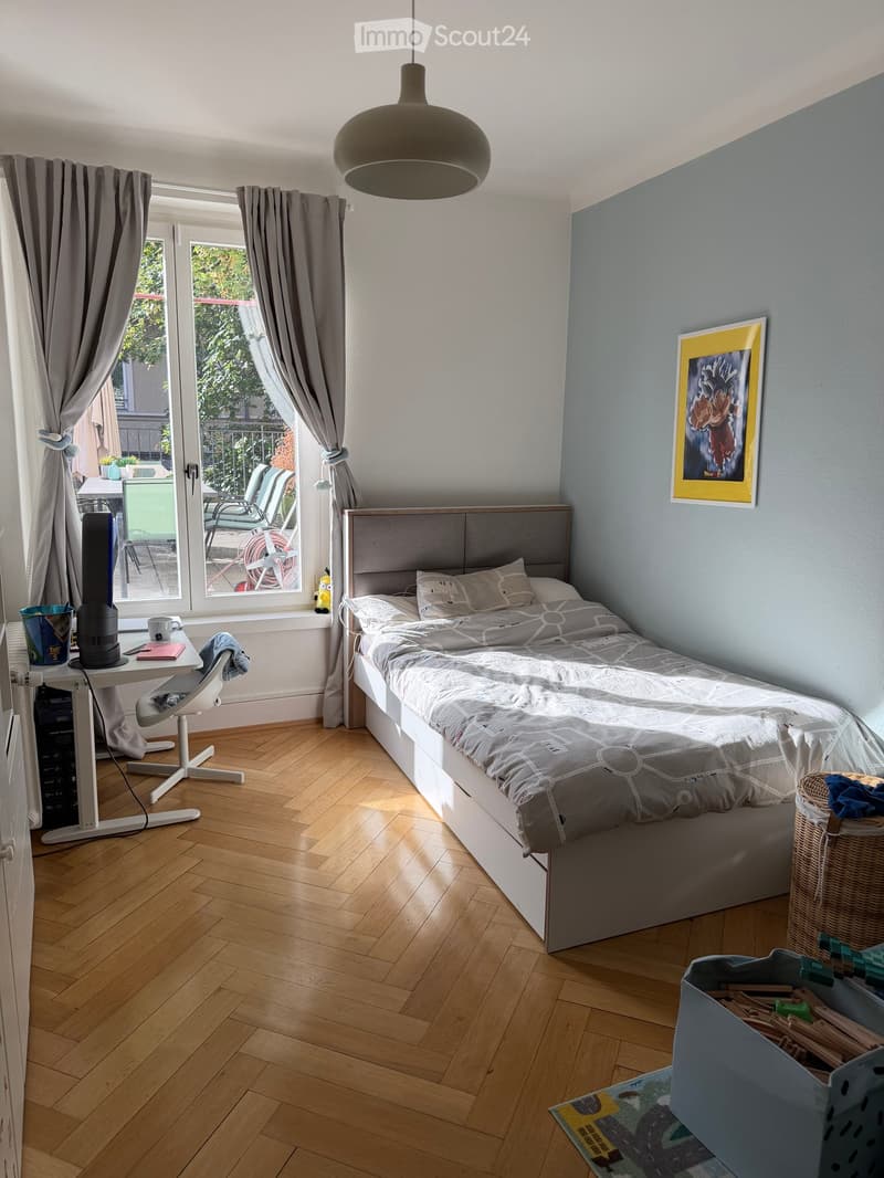 Cozy bedroom with a single bed, wooden floor, large window with curtains, desk with chair, and a hanging lamp.