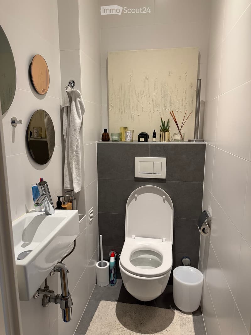 Modern bathroom with white tiles, a white sink, a toilet, a towel, a small plant, and decorative items on the counter.