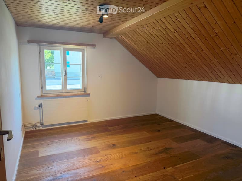 Empty attic room with wooden floors, white walls, and a skylight window. Features exposed wooden beams on the ceiling and a radiator under the window.