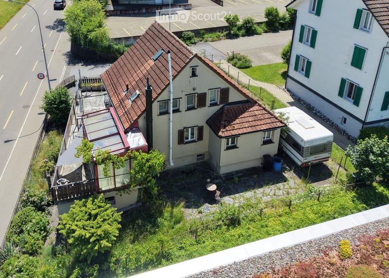 Aerial view of a two-story house with a brown tiled roof, multiple windows, and a small garden area. A camper van is parked nearby, and there is a neighboring house with green shutters visible.