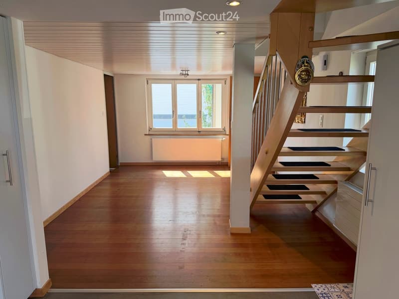 Bright hallway with wooden flooring, white walls, and a staircase leading upwards. Features include a window with blinds, a radiator, and a door on the left side.