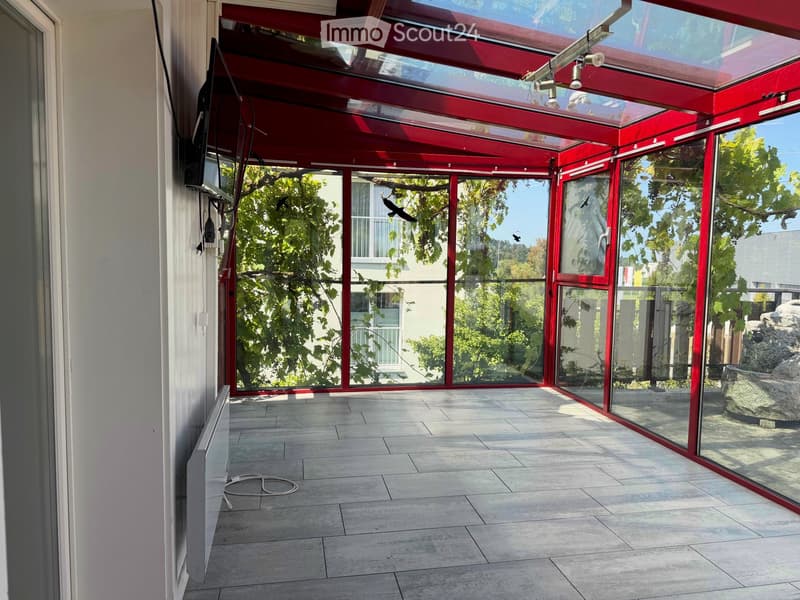 Bright, enclosed patio with red-framed glass walls and ceiling, tiled floor, and natural light. Features a stone water feature and visible outdoor greenery.