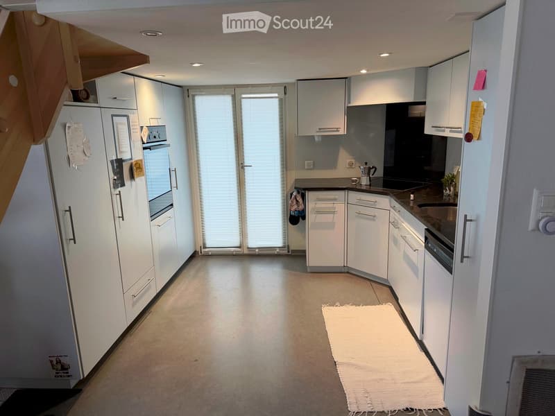 Modern kitchen with white cabinets, stainless steel appliances, and a large window. Features include a refrigerator, oven, sink, coffee maker, and a small white rug on the floor.