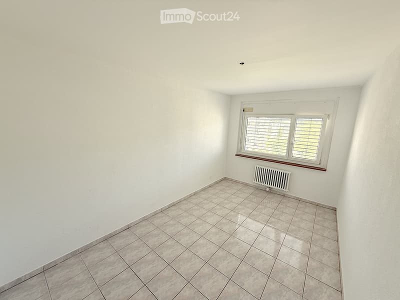 Empty room with white walls, tiled floor, and a window with blinds. A radiator is visible below the window.