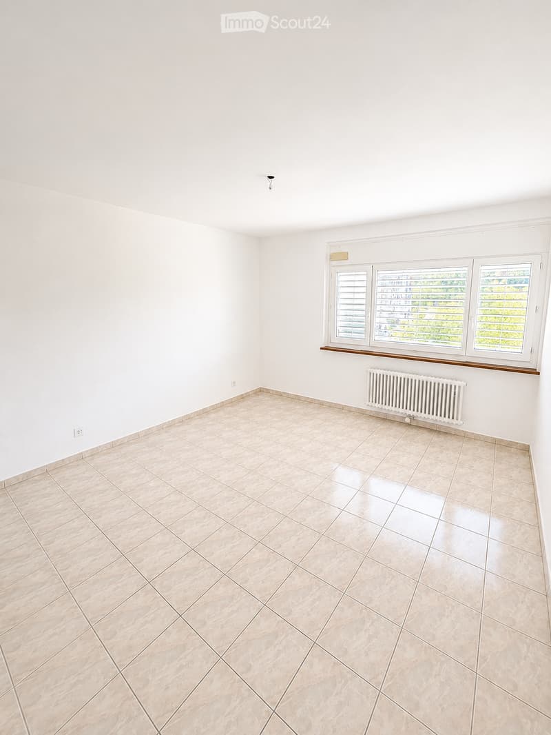Empty room with white walls, tiled floor, and three windows with blinds. A radiator is visible under the windows.