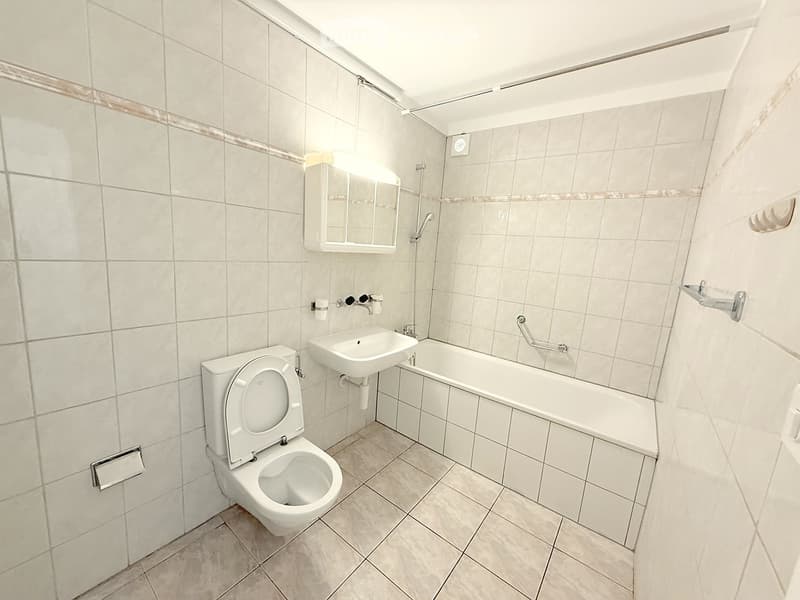 Modern bathroom with white tiled walls and floor, featuring a toilet, sink, bathtub, and a mirror above the sink.