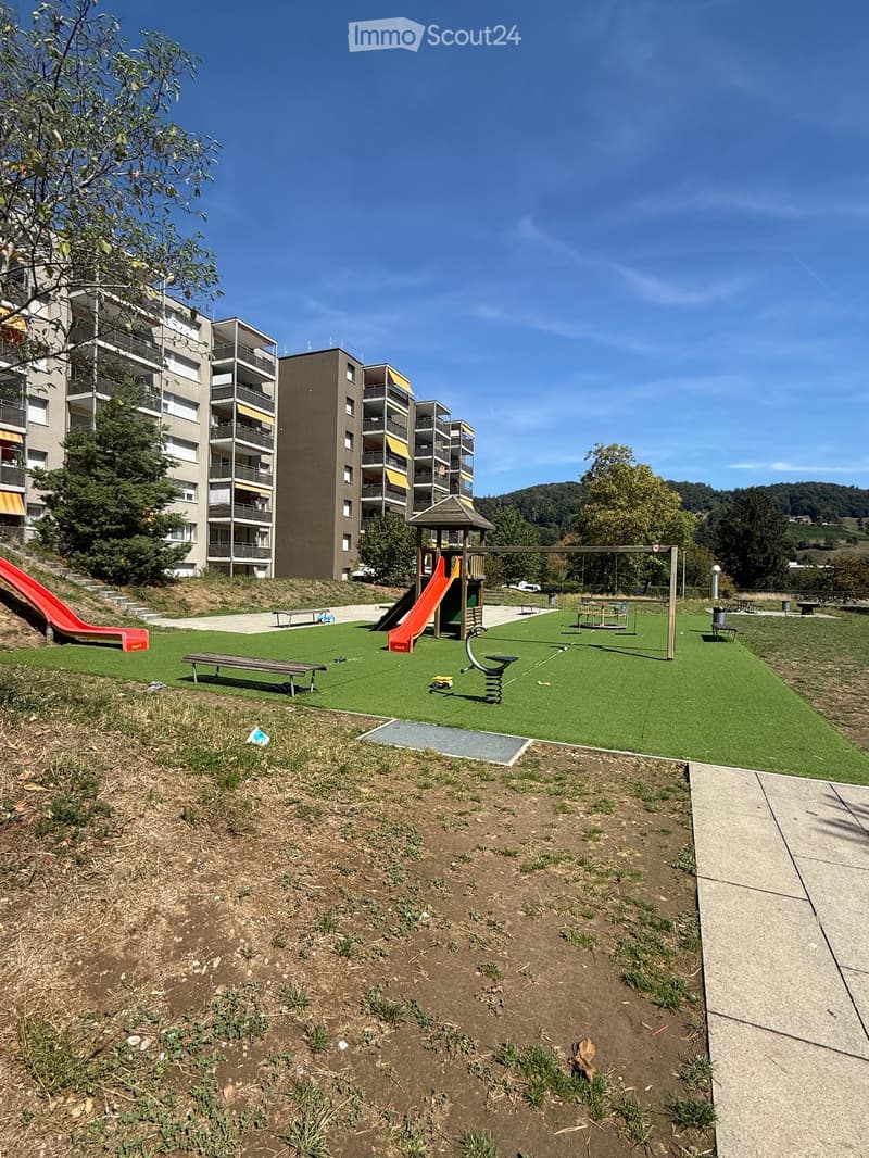 Playground area with artificial grass, swings, slides, benches, and a small gazebo, surrounded by apartment buildings and trees.