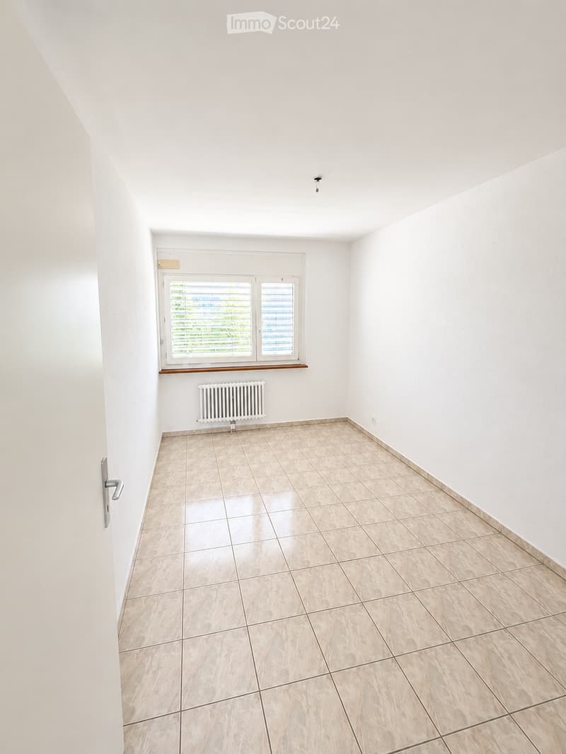 Empty room with white walls, tiled floor, window with blinds, radiator, and a door handle visible.