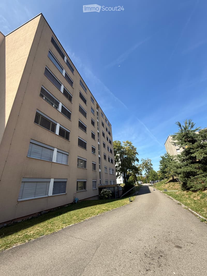Multi-story residential building with beige facade, multiple windows, and a paved pathway leading to the entrance.