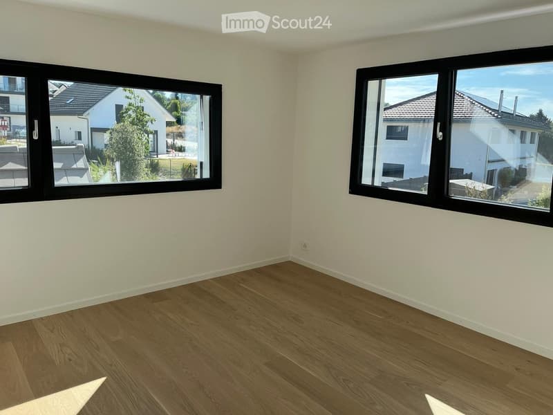 Empty room with light-colored wooden flooring and large windows offering a view of neighboring houses and greenery.