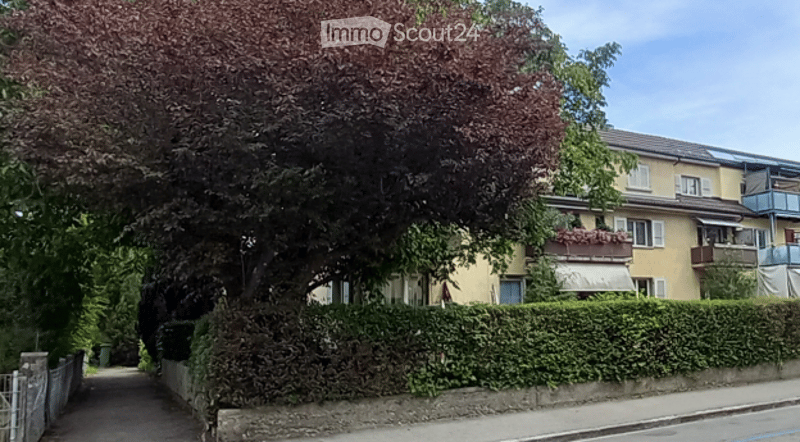 Multi-story residential building with balconies, surrounded by greenery and a well-maintained hedge.