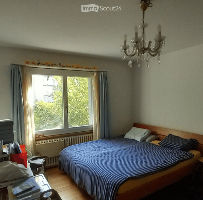 Bedroom with wooden floor, large window with curtains, bed with blue and white striped bedding, nightstand, chandelier, radiator, small table with chair.