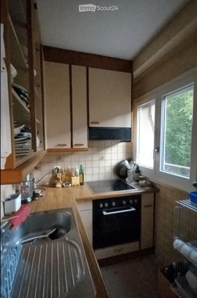 A small kitchen with wooden cabinets, a sink, an oven, and various kitchen items on the counter, including bottles and a dish rack.