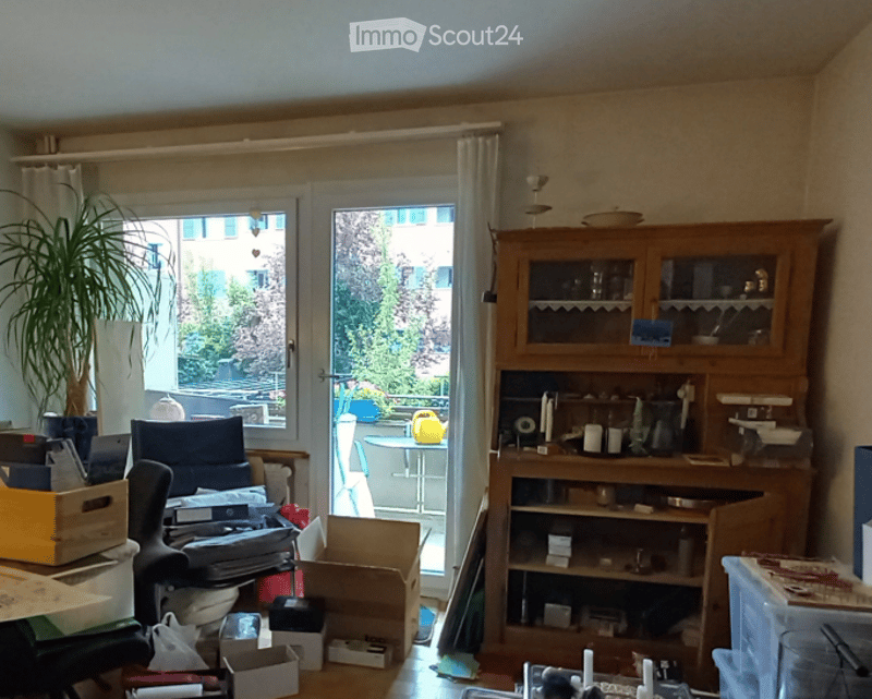 The room is furnished with a chair, a table, a wooden cupboard, and a potted plant. There is a door leading to a balcony with a view of the outside. The room appears to be in the process of being organized or unpacked, with various items and boxes scatter