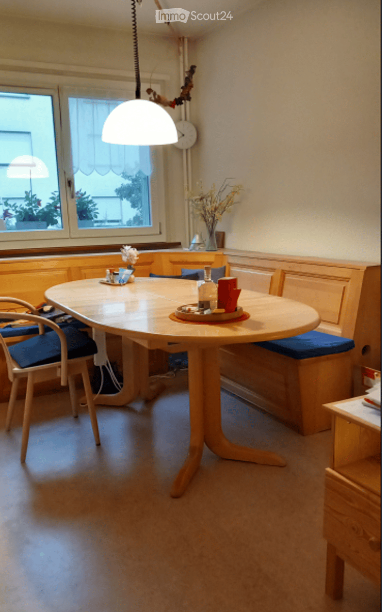 Dining area with a round wooden table, bench seating, and a single chair. A vase with yellow flowers and a cup are placed on the table. A clock is mounted on the wall above the window.