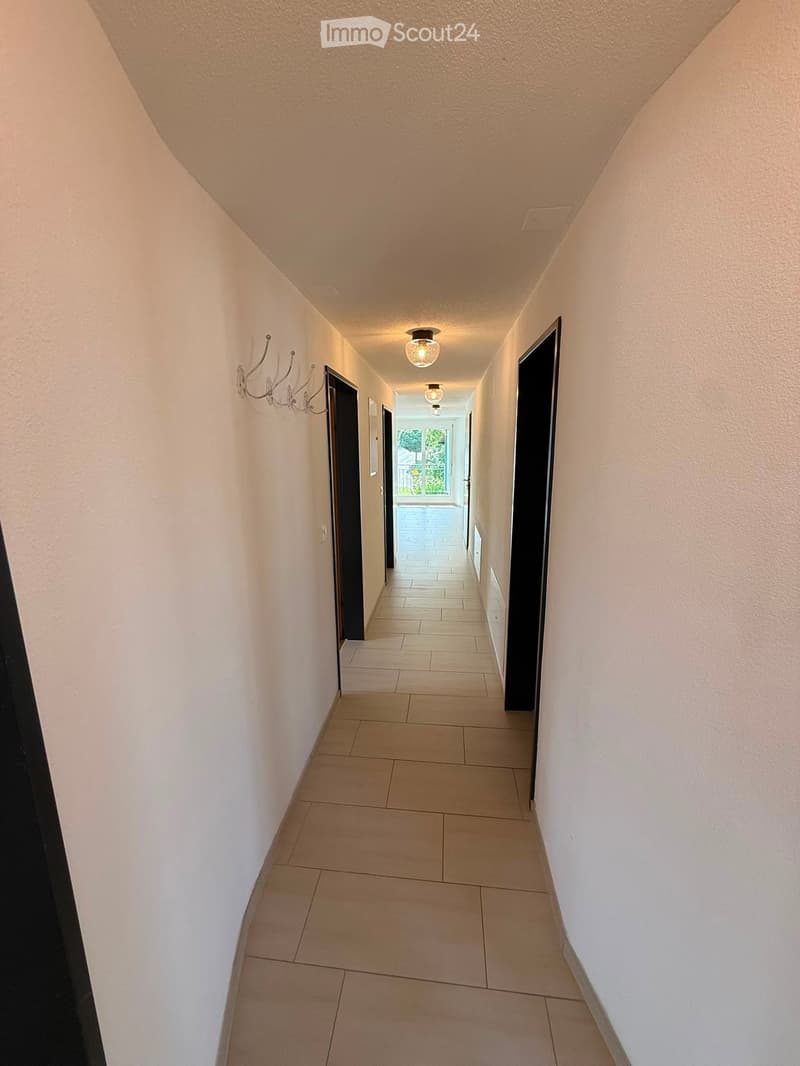 A clean, well-lit hallway with beige tiled flooring, white walls, and recessed lighting. Features include a coat rack on the left wall and several closed doors leading to other rooms.