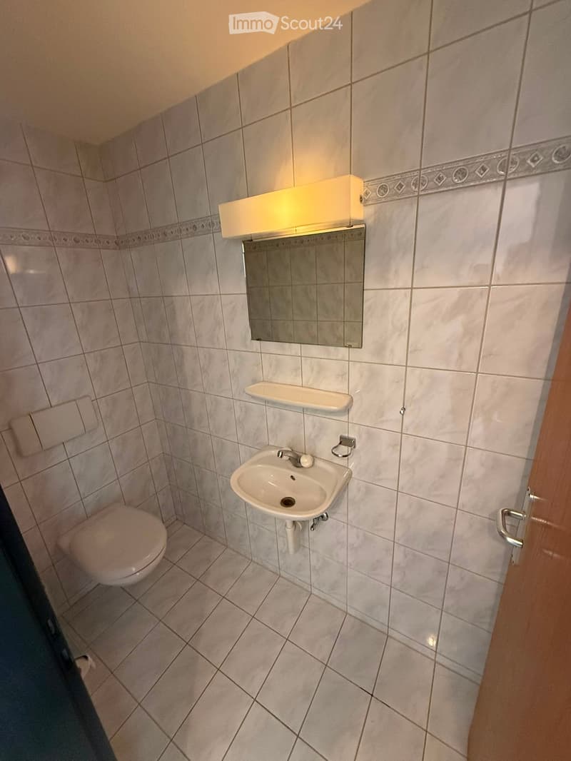Small bathroom with white tiled walls and floor, featuring a toilet, a sink with a mirror above it, and a light fixture on the wall.