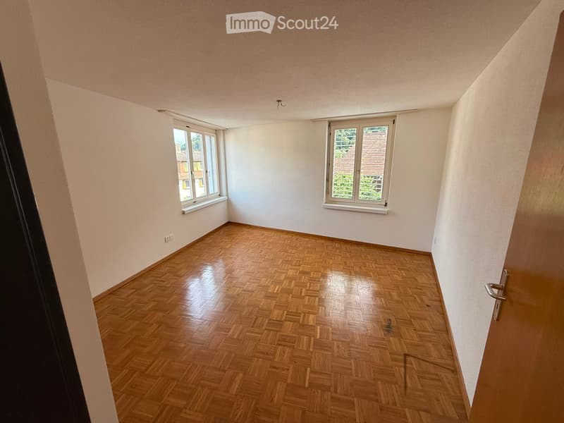 Empty room with wooden parquet flooring, white walls, and two windows offering a view of greenery outside.