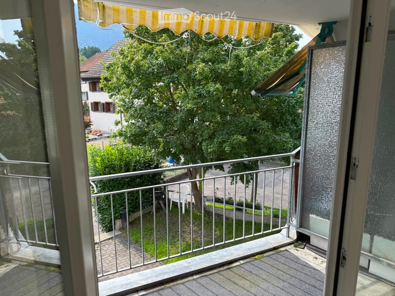 Balcony with metal railing, tiled floor, and outdoor furniture including a table and chairs, overlooking a green garden and neighboring house.