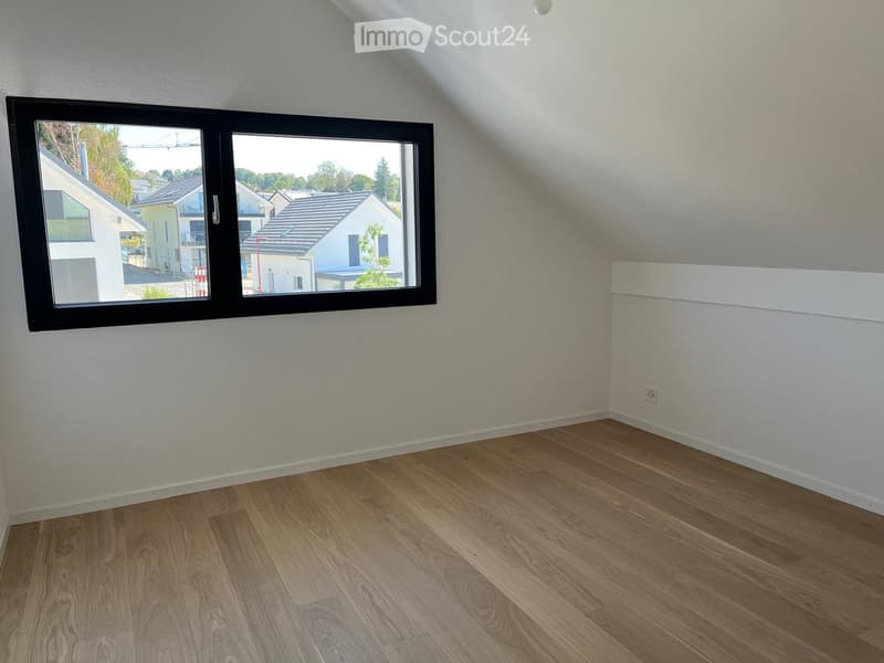 Empty attic room with wooden flooring, white walls, and a large window offering a view of the neighborhood.