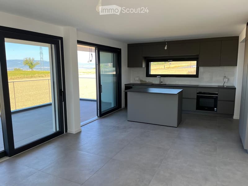 Modern kitchen with grey cabinets, a central island, and large windows overlooking a scenic outdoor area.