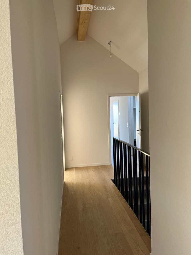 Empty hallway with wooden flooring, white walls, and a staircase leading upwards with a black railing.