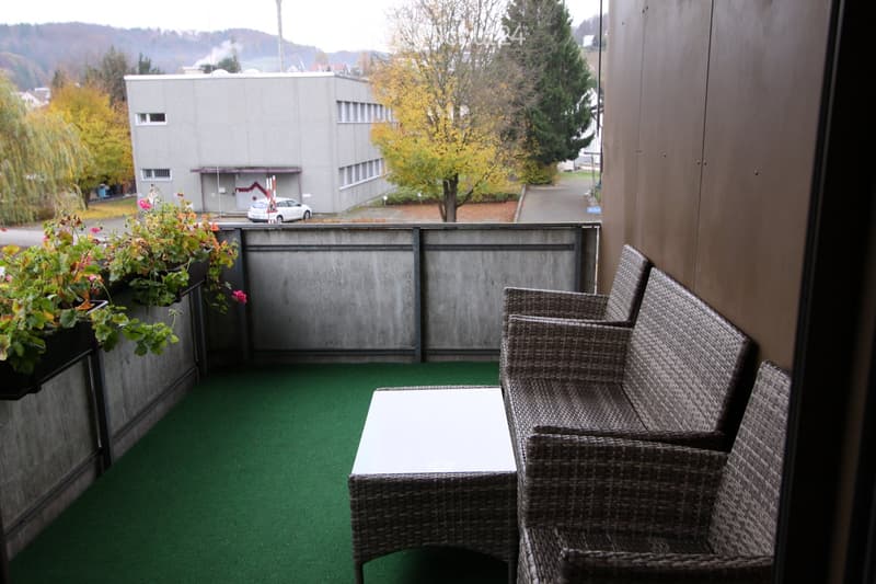 Balcony with green artificial turf, wicker furniture including a table and chairs, and potted plants on the left side.