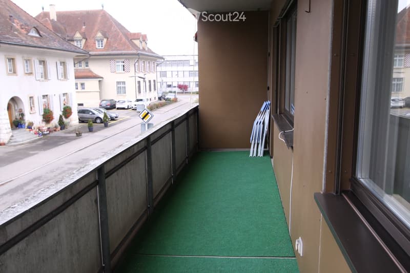 Balcony with green artificial turf flooring, metal railing, and a view of a street with parked cars and buildings.