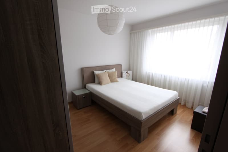Modern bedroom with a wooden bed, white bedding, two nightstands, and a large window with white curtains.