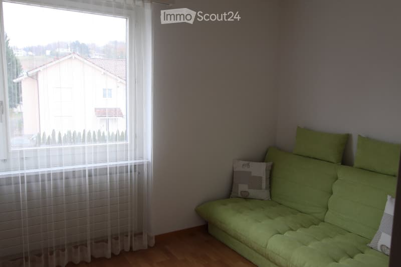 A small, cozy living space with a green sofa and matching pillows, positioned near a large window with sheer white curtains. The room has wooden flooring and a view of a neighboring house and garden outside.