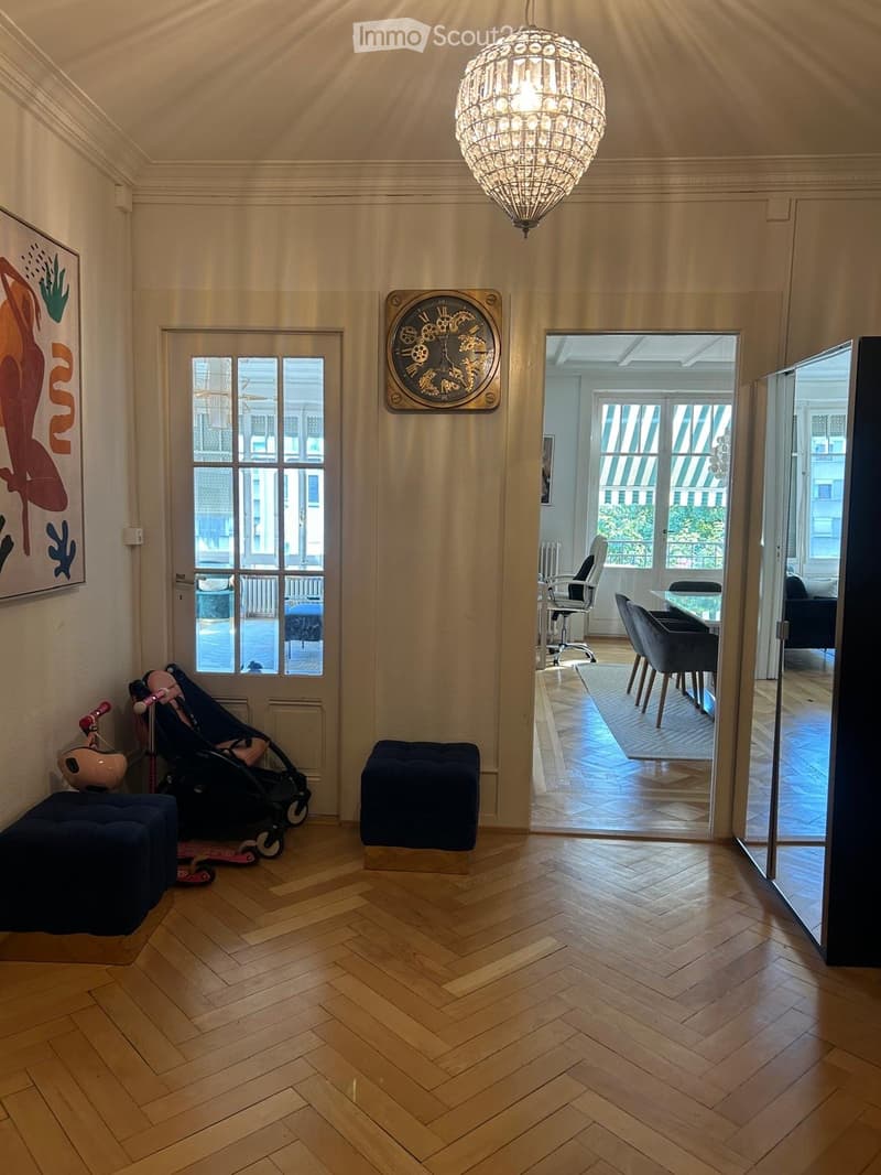 A well-lit room with wooden herringbone flooring, a large chandelier, a wall-mounted clock, and a doorway leading to another room with a dining table and chairs.