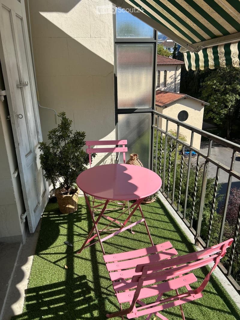 Balcony with artificial grass, pink table and chairs, potted plant, and striped awning.