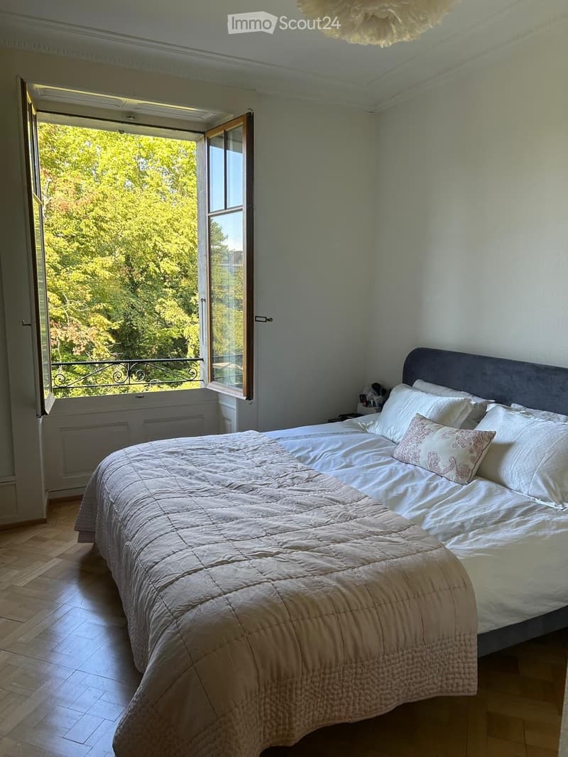 Bright bedroom with a large bed, white bedding, and a nightstand. Large windows open to a balcony with a view of greenery.