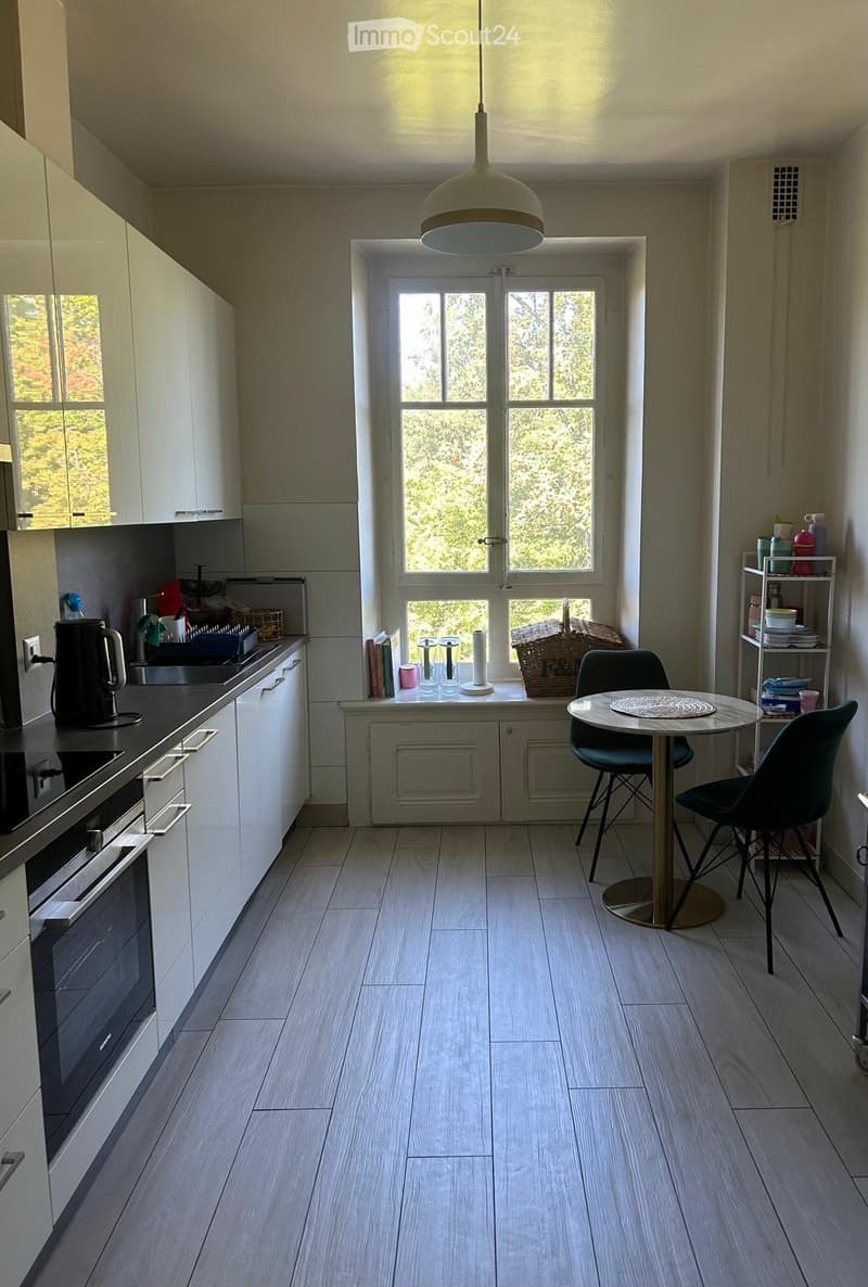 Modern kitchen with white cabinets, wooden flooring, and a round dining table with two chairs. A large window offers a view of greenery outside.