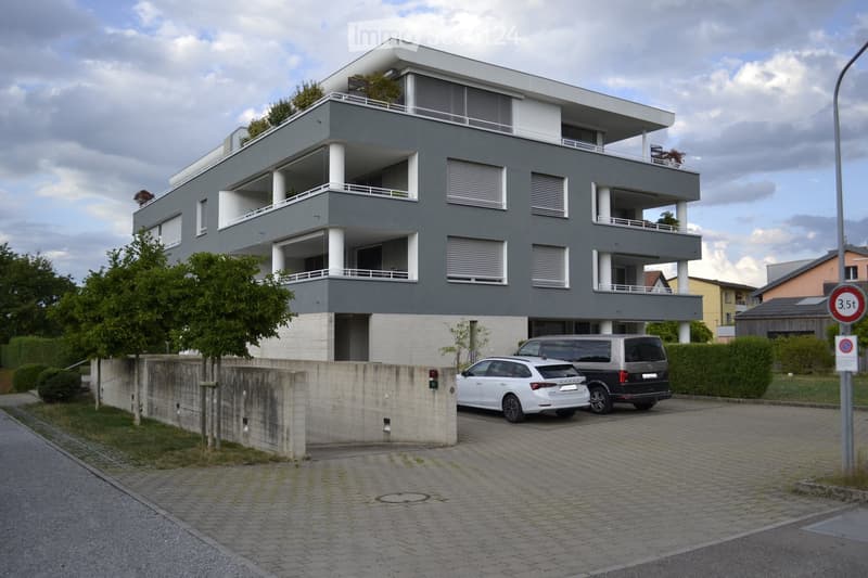 Modern three-story apartment building with grey facade, white balconies, and multiple windows. Two cars are parked in the driveway.