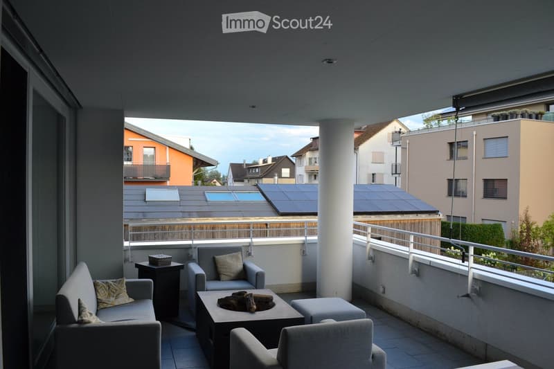 Balcony with modern furniture including a sofa, chairs, a coffee table, and a fire pit, overlooking a residential area with solar panels on rooftops.